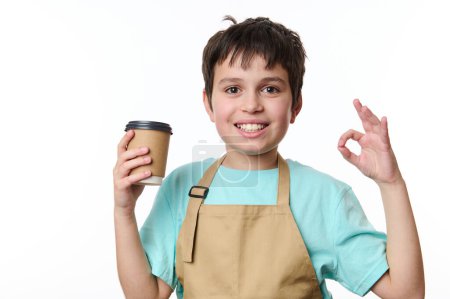 Téléchargez les photos : Portrait isolé sur fond blanc d'un garçon barman heureux adolescent montrant OK signe, la vente d'une boisson de café à emporter dans une tasse en papier éco. Boisson chaude à emporter. Espace de copie pour votre publicité - en image libre de droit