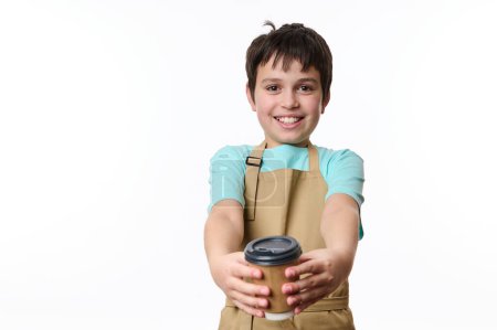 Téléchargez les photos : Adorable adolescent heureux portant un t-shirt bleu et tablier de chefs beige, souriant un sourire à la caméra, distribuant une tasse en carton écologique de café à emporter, isolé sur fond blanc - en image libre de droit