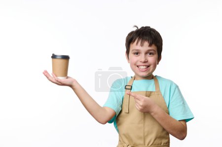 Téléchargez les photos : Beau garçon enfant portant tablier de chefs beige, souriant un sourire de dents regardant la caméra, pointant du doigt une tasse en carton écologique de café à emporter, fond blanc isolé. Espace de copie - en image libre de droit
