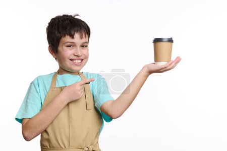 Téléchargez les photos : Un adolescent multi-ethnique portant un tablier de chef, pointant du doigt une tasse de café à emporter sur ses mains, isolé sur blanc. Espace libre pour votre publicité. Consommation d'aliments et de boissons. - en image libre de droit