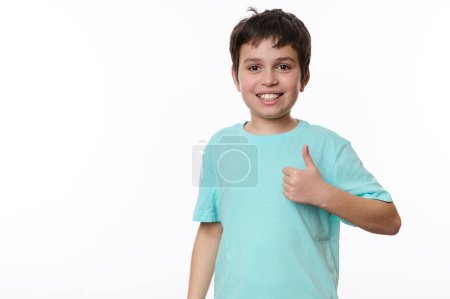 Happy Caucasian preteen child boy in blue t-shirt, a smart schoolboy shows a thumb up, cutely smiles looking at camera, isolated over white background. Free advertising space for your promotional text