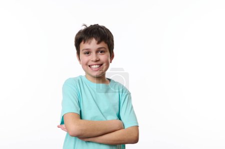 Handsome happy preteen boy in casual blue t-shirt, smiling a beautiful toothy smile, looking at camera, standing with crossed arms over white isolated background. Positive young people. Free ad space