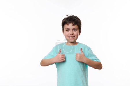 Portrait of Caucasian handsome pleasant teenage child boy in blue casual t-shirt, smiling pleasantly, looking at camera, showing thumbs up, isolated on white background with space for promotional text