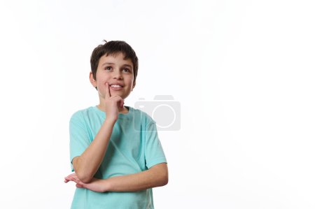 Happy teenage boy wearing turquoise t-shirt, holding his hand at chin and cutely smiling, thoughtfully looking at aside, isolated over white background with copy ad space for promotional text