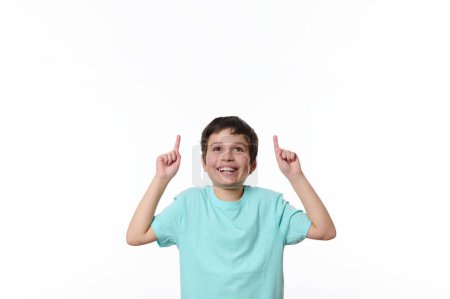 Foto de Retrato de niño preadolescente guapo caucásico agradable en camiseta casual azul, sonriendo agradablemente, mirando hacia arriba, señalando con el dedo el espacio de copia para su texto publicitario, aislado sobre fondo blanco - Imagen libre de derechos