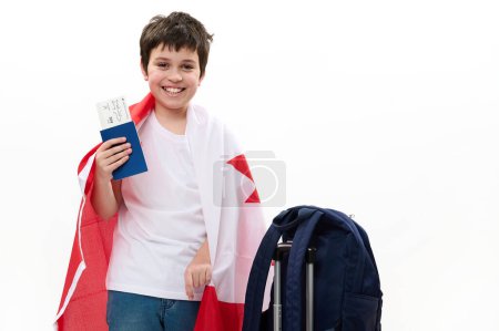 Foto de Multiétnico feliz adolescente inteligente en camiseta casual y vaqueros azules, sonriendo, mirando a la cámara, posando con bandera canadiense, tarjeta de embarque y mochila sobre fondo blanco. Espacio libre para publicidad - Imagen libre de derechos