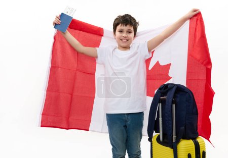 Foto de Un adolescente caucásico inteligente con camiseta blanca y vaqueros azules, con mochila, maleta y tarjeta de embarque, con bandera canadiense, viaja a Canadá para estudiar intercambio de experiencias. Inmigración - Imagen libre de derechos