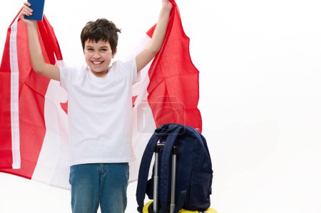 Foto de Jovencito adolescente americano vestido con camiseta casual y vaqueros azules, sonriendo alegremente, posando con bandera canadiense, tarjeta de embarque y equipaje aislado sobre fondo blanco. Espacio publicitario - Imagen libre de derechos