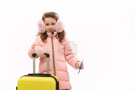 Téléchargez les photos : Portrait d'une jolie petite fille caucasienne portant des cache-oreilles roses, une veste en duvet et des mitaines en laine, souriant en regardant la caméra, posant avec une valise et une carte d'embarquement, isolée sur fond blanc - en image libre de droit