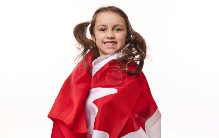 Foto de Niña encantadora americana, ciudadana canadiense envolviendo la bandera de Canadá, sonriendo una hermosa sonrisa dentada mirando a la cámara, aislada sobre fondo blanco. Copia el espacio publicitario. Día del Dominio. 1 de julio - Imagen libre de derechos