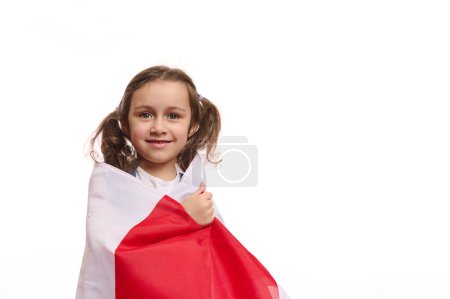 Foto de Niña europea con dos lindas colas de caballo envolviendo la bandera de Polonia, sonriendo alegre sonrisa dentada mirando a la cámara, aislada sobre fondo blanco. Celebración del Día de la Independencia de Polonia, 11 de noviembre - Imagen libre de derechos