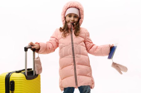 Téléchargez les photos : Charmante petite voyageuse vêtue d'un duvet rose chaud, debout près d'une valise jaune avec une carte d'embarquement à la main, allant pour une escapade de week-end, isolée sur fond blanc. Vacances d'hiver - en image libre de droit