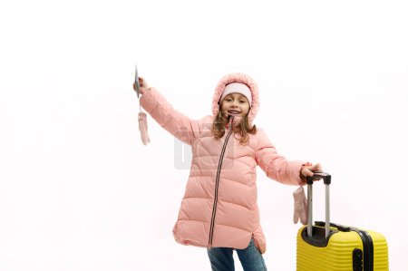 Téléchargez les photos : Belle petite fille en manteau d'hiver rose chaud, avec billet d'avion, carte d'embarquement et valise jaune, vagues bonjour, regardant la caméra, isolé sur fond blanc. Escapade de week-end. Tourisme de voyage concept - en image libre de droit