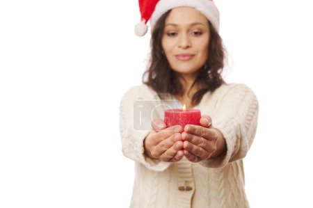 Téléchargez les photos : Détails : bougie de Noël rouge allumée dans les mains d'une belle femme brune floue, portant un chapeau de Père Noël et un pull beige chaud, isolé sur fond blanc. Noëlmastime. Bonne année. Lumière des bougies - en image libre de droit