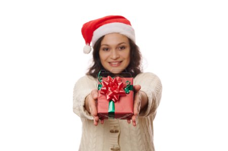 Téléchargez les photos : Focus sélectif sur un joli cadeau de Noël, enveloppé dans du papier d'emballage rouge et noué avec un ruban vert, dans les mains d'une belle femme souriante floue, isolée sur fond blanc. Nouvel An - en image libre de droit