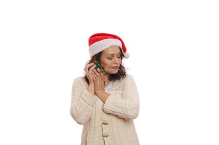 Téléchargez les photos : Jolie femme multi-ethnique sereine portant le chapeau de Père Noël, embrassant doucement son cadeau de Noël, posant les yeux fermés, isolée sur fond blanc avec un espace publicitaire de copie. Concept du Nouvel An - en image libre de droit