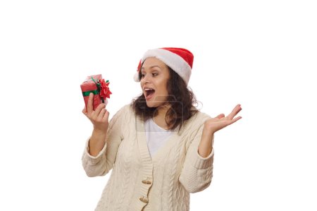 Téléchargez les photos : Portrait isolé sur fond blanc d'une jolie femme portant le chapeau du Père Noël, se réjouissant du cadeau de Noël entre ses mains. Noël, préparatifs du Nouvel An. Profiter de l'ambiance festive des prochaines vacances d'hiver - en image libre de droit