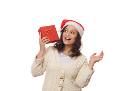 Téléchargez les photos : Jolie jolie femme au chapeau de Père Noël, secouant une belle boîte cadeau, devinant ce qu'elle contient, isolée sur fond blanc. Il est temps de déballer les cadeaux de Noël et du Nouvel An. Joyeuses vacances d'hiver - en image libre de droit