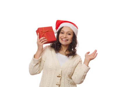 Téléchargez les photos : Belle femme latino-américaine en chapeau de Père Noël avec boîte cadeau de Noël, souriant un sourire de dents regardant la caméra, isolé sur fond blanc. Il est temps de déballer les cadeaux de Noël et du Nouvel An. Jour de boxe - en image libre de droit