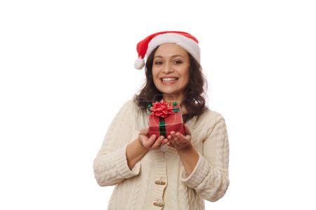 Téléchargez les photos : Brune multi-ethnique, une heureuse femme d'âge moyen au chapeau de Père Noël et pull beige, montrant un cadeau de Noël en papier d'emballage rouge et arc, souriant mignon, regardant la caméra, sur fond blanc - en image libre de droit