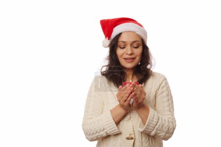 Téléchargez les photos : Femme brune au chapeau de Père Noël, en prévision des vacances d'hiver, faire vœu tout en tenant une bougie de Noël dans ses mains, isolé sur fond blanc avec espace publicitaire copie. Nouvel An. atmosphère de Noël - en image libre de droit