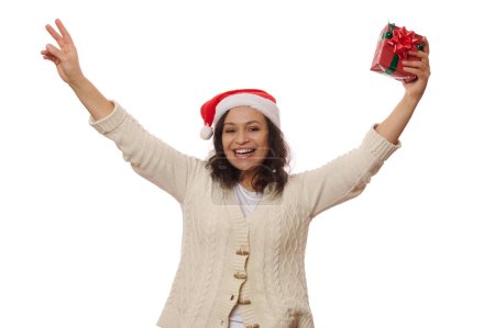 Téléchargez les photos : Belle femme afro-américaine ravie en chapeau de Père Noël, tient un cadeau de Noël dans ses mains, souriant joliment en regardant la caméra, isolé sur fond blanc. Fête du Nouvel An et concept de Noël - en image libre de droit