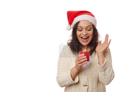 Téléchargez les photos : Hispanique surpris belle femme dans le chapeau du Père Noël et pull beige chaud, en regardant une bougie rouge allumée sur fond blanc. Ambiance de Noël. Espace publicitaire gratuit pour votre texte promotionnel - en image libre de droit