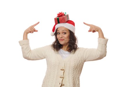 Téléchargez les photos : Femme ravie de porter le chapeau de Père Noël, regardant la caméra et pointant le cadeau de Noël sur sa tête, isolée sur fond blanc avec de l'espace publicitaire. Nouvel An, Noël et vacances d'hiver concept - en image libre de droit