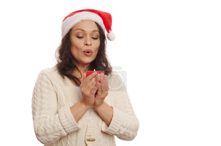 Téléchargez les photos : Charmante brune, femme latino-américaine au chapeau de Père Noël et pull chaud beige, soufflant bougie tout en faisant vœu chéri pour Noël, isolé sur fond blanc avec espace publicitaire gratuit - en image libre de droit