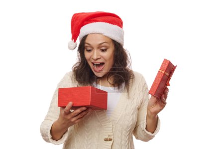 Téléchargez les photos : Belle femme afro-américaine au chapeau de Père Noël, ouvre une boîte cadeau avec une surprise, sur fond blanc. Noël Nouvel An préparatifs. Concept de Boxing Day. Bonne ambiance festive des vacances d'hiver - en image libre de droit