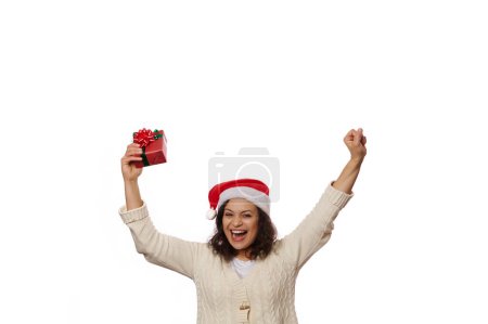 Téléchargez les photos : Jolie jolie femme au chapeau de Père Noël serrant les poings, se réjouissant du cadeau de Noël entre ses mains, isolée sur fond blanc avec de l'espace pour le texte promotionnel. Profiter des prochaines vacances d'hiver - en image libre de droit
