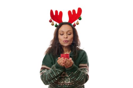 Téléchargez les photos : Belle femme multi-ethnique aux cheveux bruns, portant un cerceau en bois de cerf et un pull en laine verte à motifs de Noël, soufflant une bougie rouge sur fond blanc. Joyeux Noël. Bonne année concept. Espace publicitaire - en image libre de droit