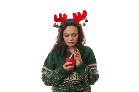 Téléchargez les photos : Belle femme aux cheveux foncés, portant un pull vert chaud à motifs de Noël et un cerceau de bois de cerf, tenant une bougie rouge allumée, isolée sur fond blanc avec un espace publicitaire gratuit. Joyeux Noël - en image libre de droit