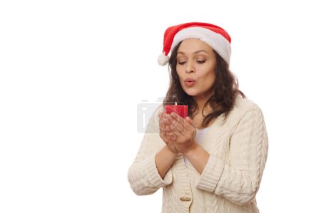 Téléchargez les photos : Amérique latine ravissante jolie femme portant le chapeau du Père Noël, soufflant une bougie rouge, isolé sur fond blanc. Portrait d'une charmante brune profitant des prochaines vacances d'hiver. Noël - en image libre de droit