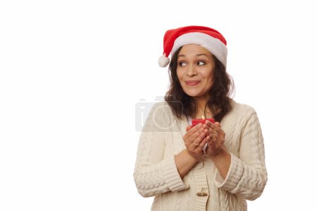 Téléchargez les photos : Belle femme brune latino-américaine, portant un chapeau de Père Noël et un pull chaud beige, regardant de côté l'espace publicitaire de copie sur fond blanc, posant avec une bougie de Noël allumée en rouge dans ses mains. - en image libre de droit