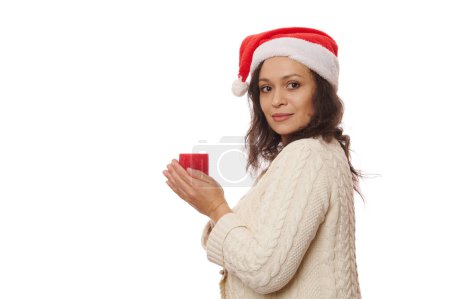 Téléchargez les photos : Portrait latéral d'une jolie femme latino-américaine au chapeau de Père Noël et au pull beige, tenant une bougie de Noël rouge allumée et regardant la caméra à travers son épaule, isolée sur fond blanc. Espace de copie - en image libre de droit