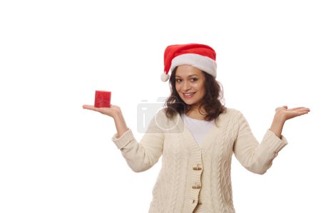 Téléchargez les photos : Belle femme multi-ethnique portant le chapeau de Père Noël, souriant d'un sourire de dents, tenant l'espace publicitaire de copie imaginaire sur une main paume vers le haut et une bougie allumée rouge sur l'autre, isolé sur fond blanc - en image libre de droit