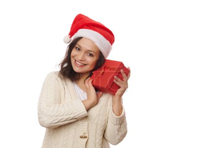 Téléchargez les photos : Jolie femme multi-ethnique au chapeau de Père Noël, embrassant doucement une boîte-cadeau avec une surprise heureuse pour Noël, et souriant mignon un sourire de dent regardant la caméra, isolé sur fond blanc. Copier l'espace publicitaire - en image libre de droit
