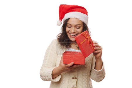 Téléchargez les photos : Joyeux ravi jolie femme dans le chapeau de Père Noël, exprimant des émotions positives tout en déballant une jolie boîte-cadeau avec surprise pour Noël, isolé sur fond blanc. Préparatifs de Noël et du Nouvel An - en image libre de droit