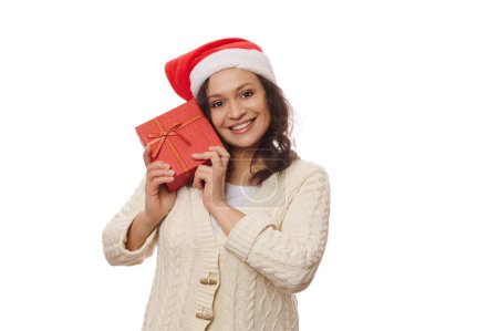 Téléchargez les photos : Jolie femme multi-ethnique d'âge moyen portant un chapeau de Père Noël, tenant une boîte cadeau de Noël près de son visage, souriant à un sourire joyeux en regardant la caméra, isolé sur fond blanc, copier l'espace publicitaire - en image libre de droit