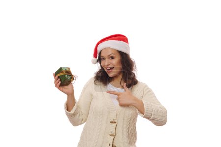 Téléchargez les photos : Jolie femme afro-américaine au chapeau de Père Noël et pull beige, pointant vers un cadeau de Noël heureux, souriant en regardant la caméra, isolé sur fond blanc. Le jour de la boxe. Temps d'ouvrir des boîtes-cadeaux - en image libre de droit