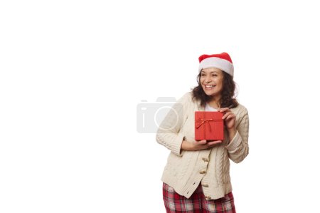 Téléchargez les photos : Portrait taille haute sur fond blanc d'une jolie femme positive multi-ethnique portant le chapeau de Père Noël, souriant joyeusement regardant de côté l'espace publicitaire de copie, posant avec la boîte-cadeau avec le cadeau heureux de Noël - en image libre de droit