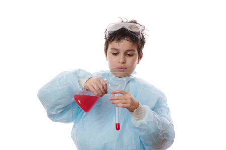 Téléchargez les photos : Un adolescent concentré, un écolier, un jeune scientifique chimiste en blouse de laboratoire, verse de la substance chimique liquide rouge à travers un entonnoir de verre dans une éprouvette sur fond blanc. Apprendre la chimie. Retour à l'école - en image libre de droit