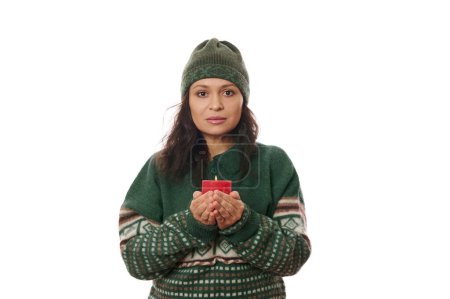 Téléchargez les photos : Belle femme aux cheveux bruns confiante, portant un chapeau chaud en laine et un pull vert tricoté à motifs de Noël, tenant une bougie rouge avec flamme et confidente regardant la caméra, isolée sur fond blanc - en image libre de droit