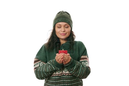 Téléchargez les photos : Belle femme latino-américaine, portant un chapeau chaud en laine et un pull vert tricoté à motifs de Noël, les yeux fermés, tenant une bougie allumée rouge et faisant un vœu chéri pour Noël, fond blanc - en image libre de droit