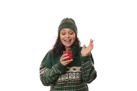 Téléchargez les photos : Femme heureuse portant un chapeau en laine chaude et un pull vert à motifs de Noël, se réjouit d'une bougie rouge allumée dans ses mains, souriant d'un sourire joyeux à une flamme de feu, isolé sur fond blanc - en image libre de droit
