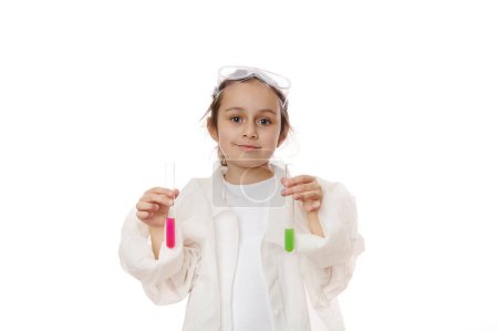Téléchargez les photos : Jolie petite fille, future chimiste scientifique en blouse de laboratoire, sourit en regardant la caméra, tenant des éprouvettes avec des produits chimiques liquides, isolées sur fond blanc. Les enfants apprennent la chimie. Concept d'éducation - en image libre de droit