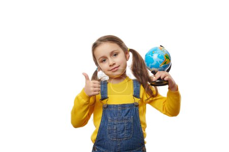 Téléchargez les photos : Charmante petite fille, un enfant conscient, tenant un globe et montrant pouce levé regardant la caméra, profitant de cours de géographie, isolé sur fond blanc avec de l'espace publicitaire. Jour de la Terre. Sauver la planète - en image libre de droit