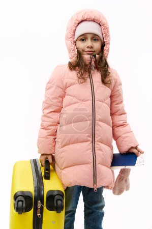 Téléchargez les photos : Adorable petite fille caucasienne portant un duvet rose, allant pour les vacances d'hiver, voyageant à l'étranger, tenant une carte d'embarquement et une valise jaune, souriant en regardant la caméra sur fond blanc. Espace de copie - en image libre de droit