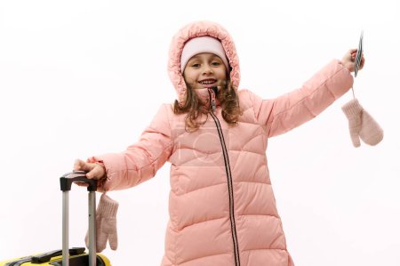 Téléchargez les photos : Joyeuse petite fille dans des vêtements chauds et duveteux, allant pour les vacances d'hiver, levant les mains dans l'excitation, attendant le voyage aérien à venir, posant avec une carte d'embarquement et une valise sur fond blanc - en image libre de droit
